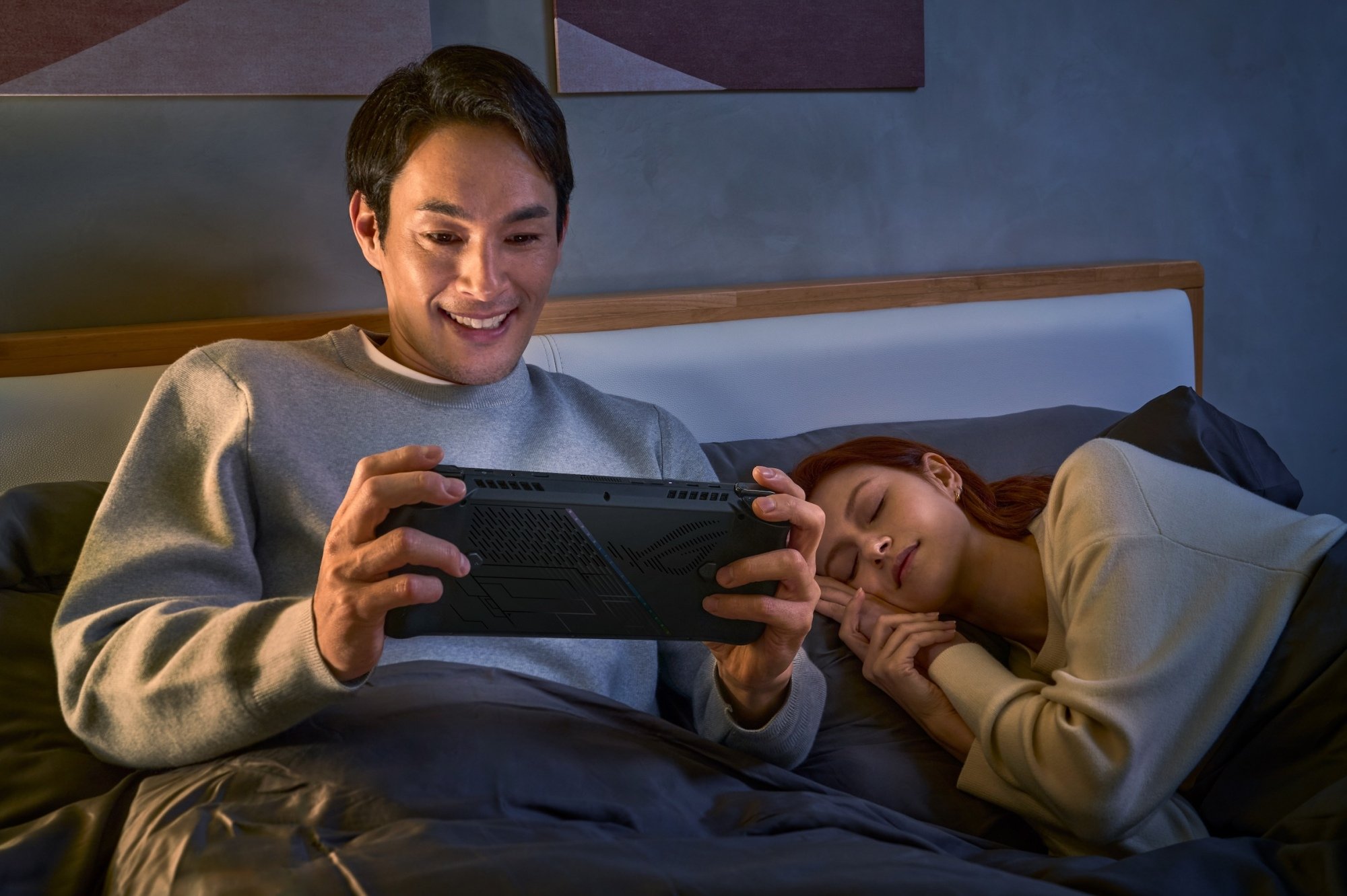 Man in bed holding Asus ROG Ally X while woman sleeps next to him