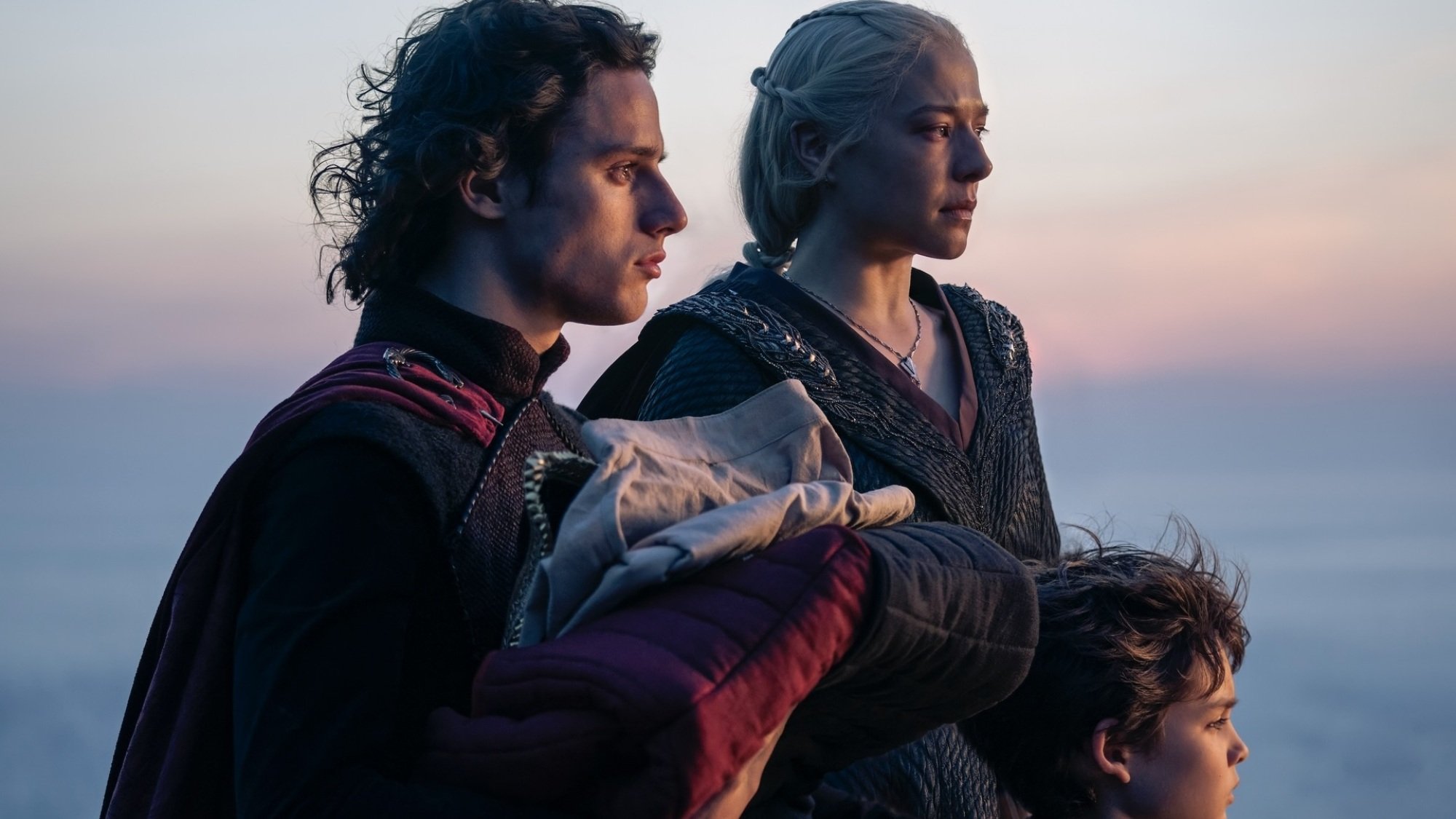 Jacaerys, Rhaenyra, and Joffrey stand together with the sun setting over the ocean behind them.