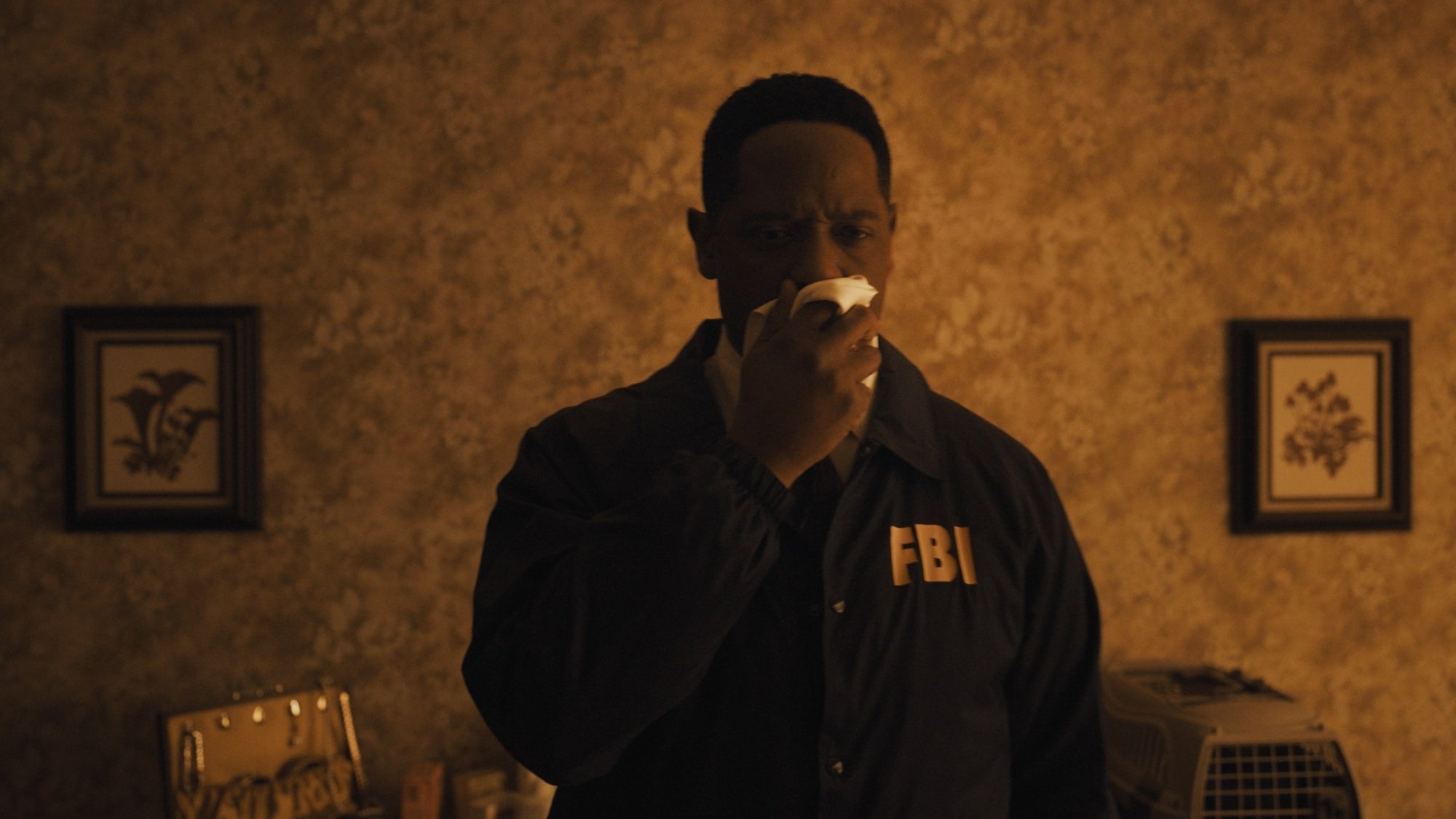 A man in an FBI jacket covers his nose while in a bedroom.