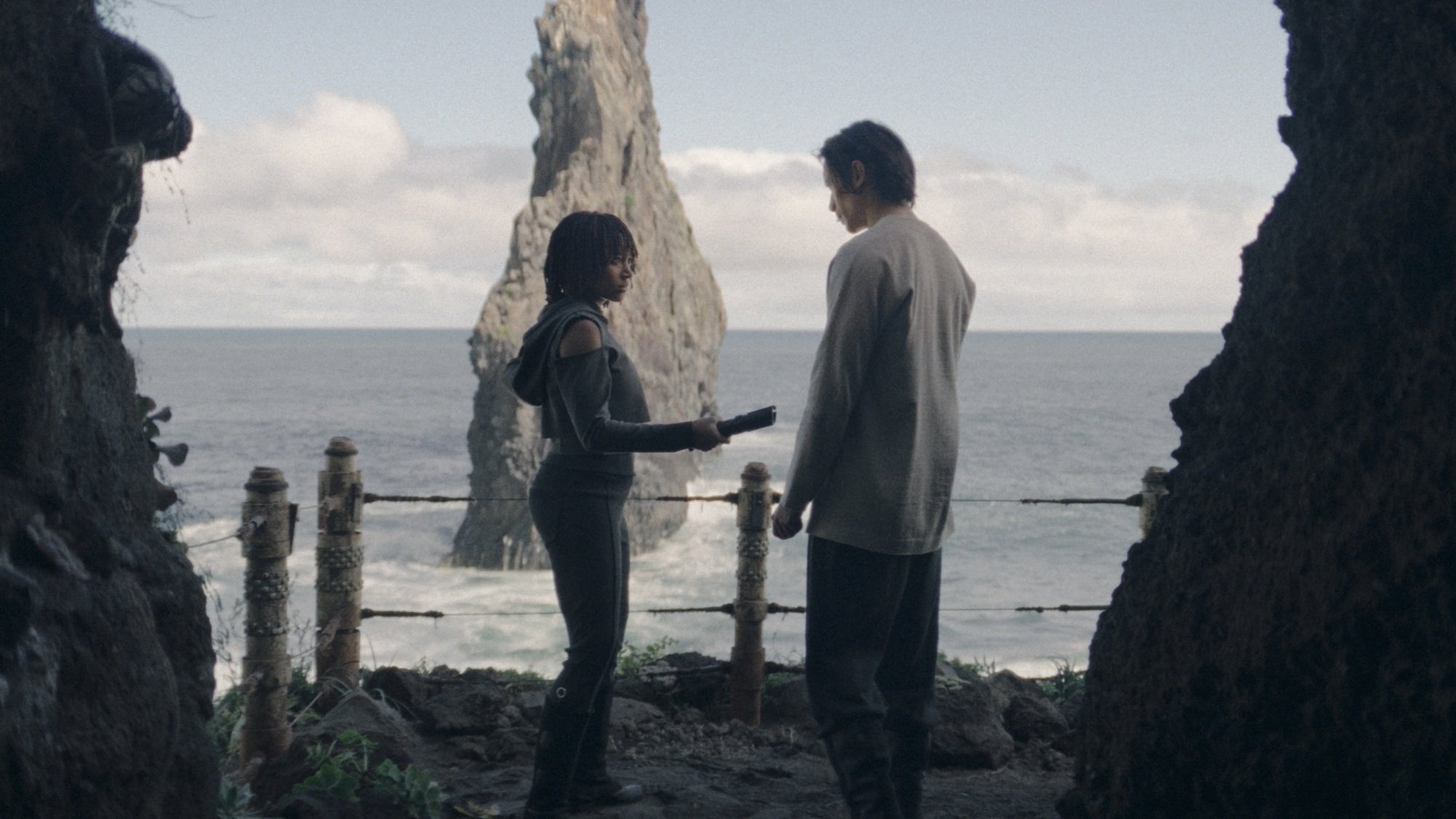 Osha holds an unlit lightsaber between her and the Stranger; the two stand in a cave mouth overlooking the ocean.