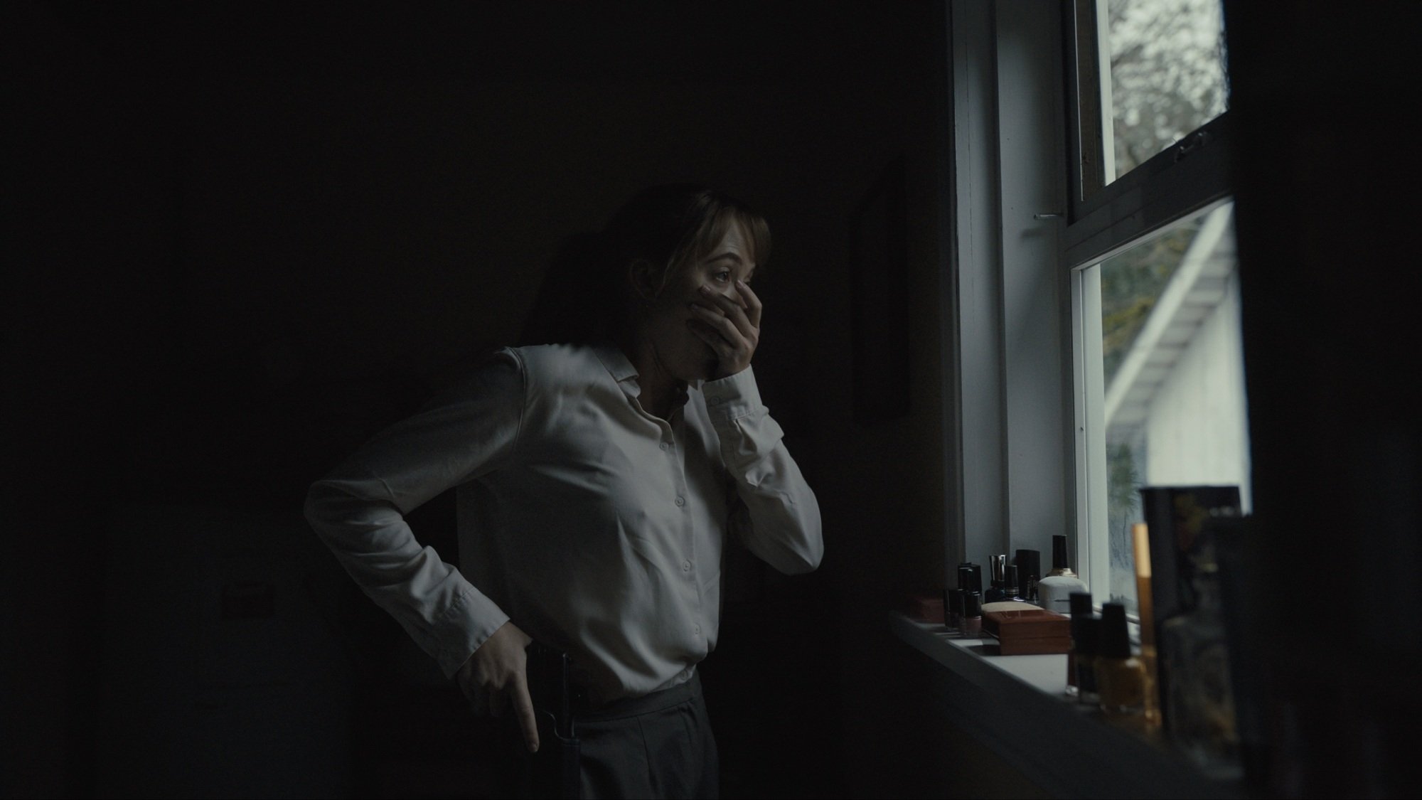 A woman in a white button-up shirt covers her mouth in horror while looking out a window.