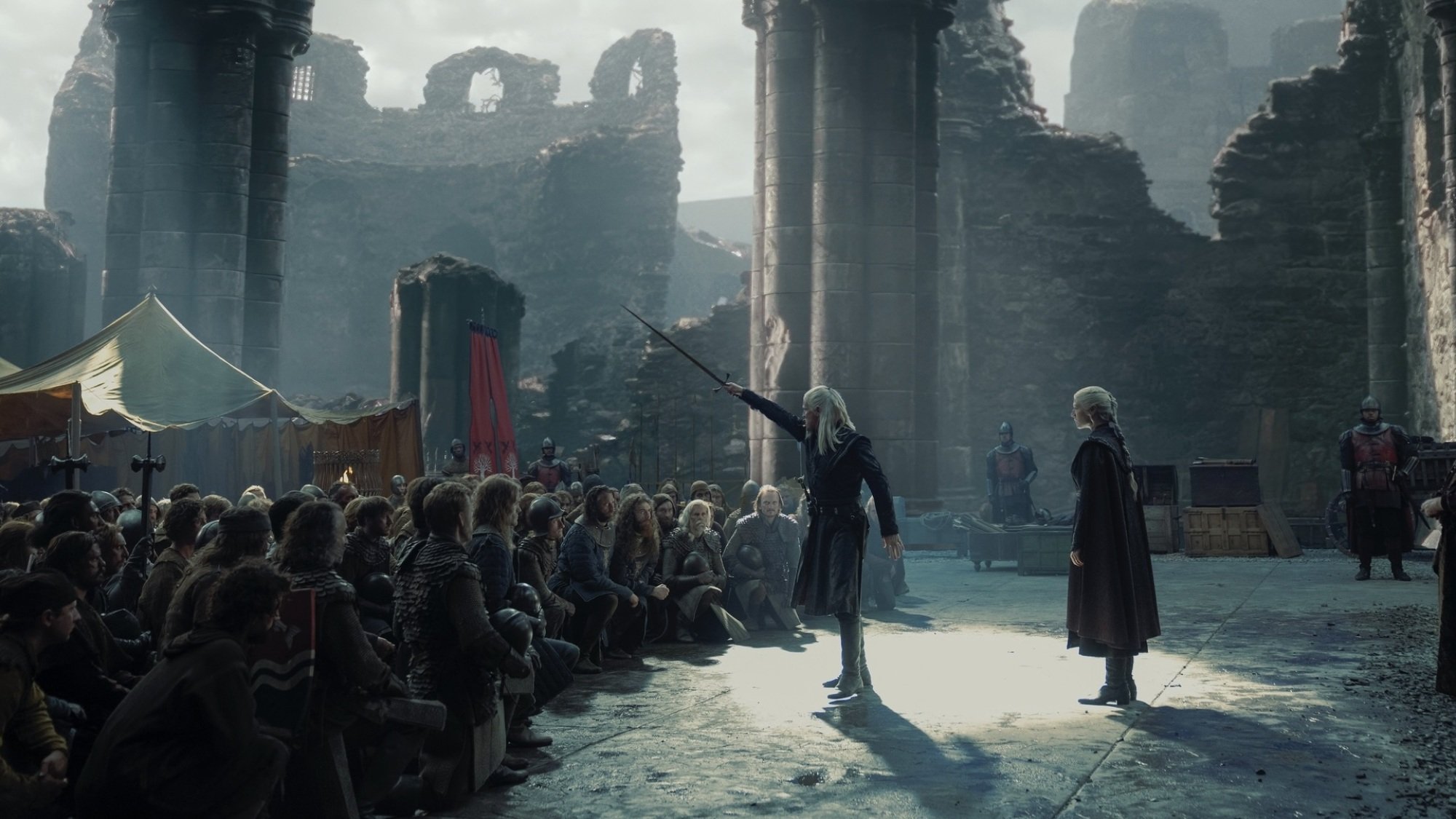 Daemon Targaryen raises his sword at the head of a kneeling army in Harrenhal; Rhaenyra Targaryen stands behind him.