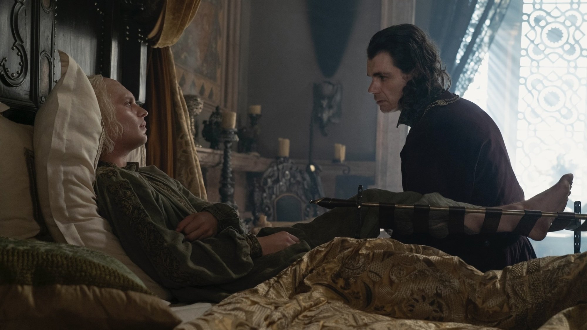 Aegon Targaryen lies wounded in bed, with his left leg propped up in a brace; Larys Strong sits beside him on the edge of the bed.