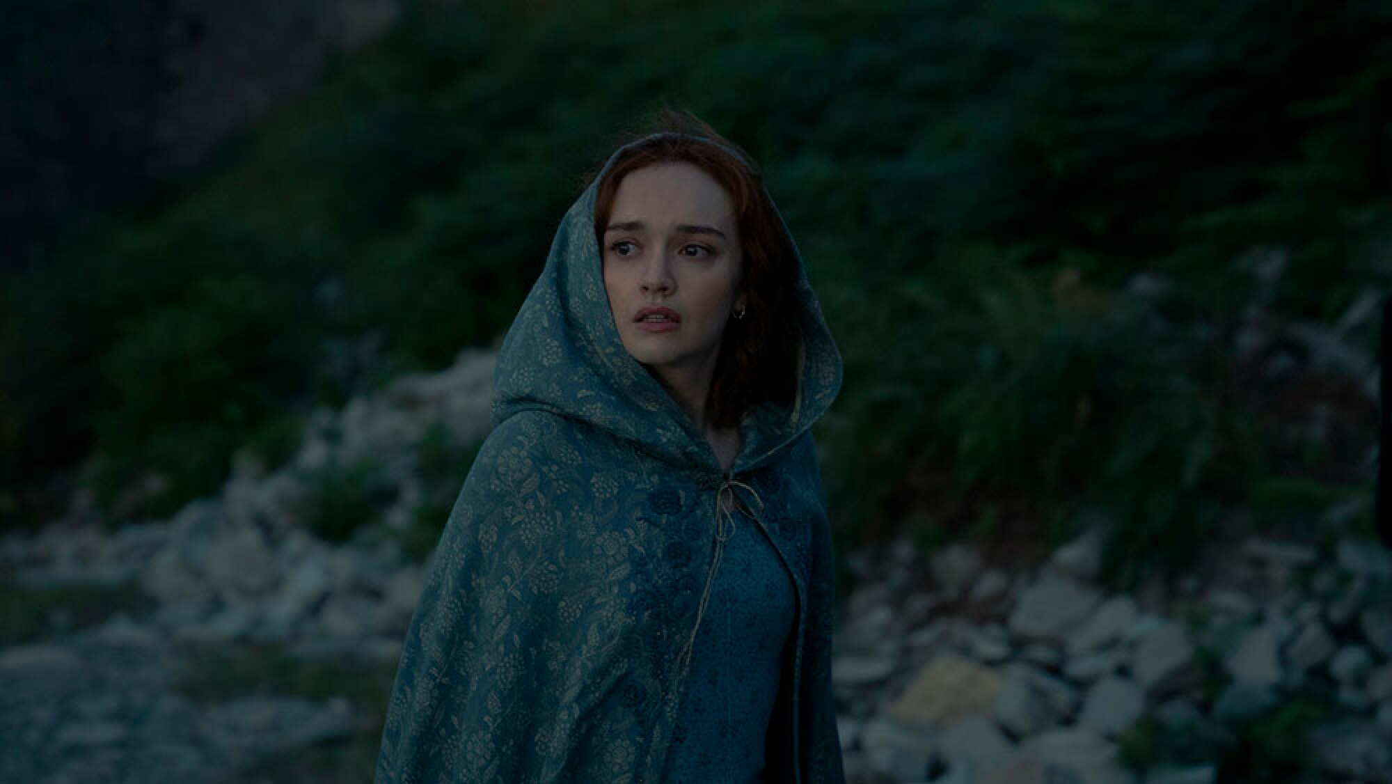 A women in a blue cloak walks through the darkness outside.