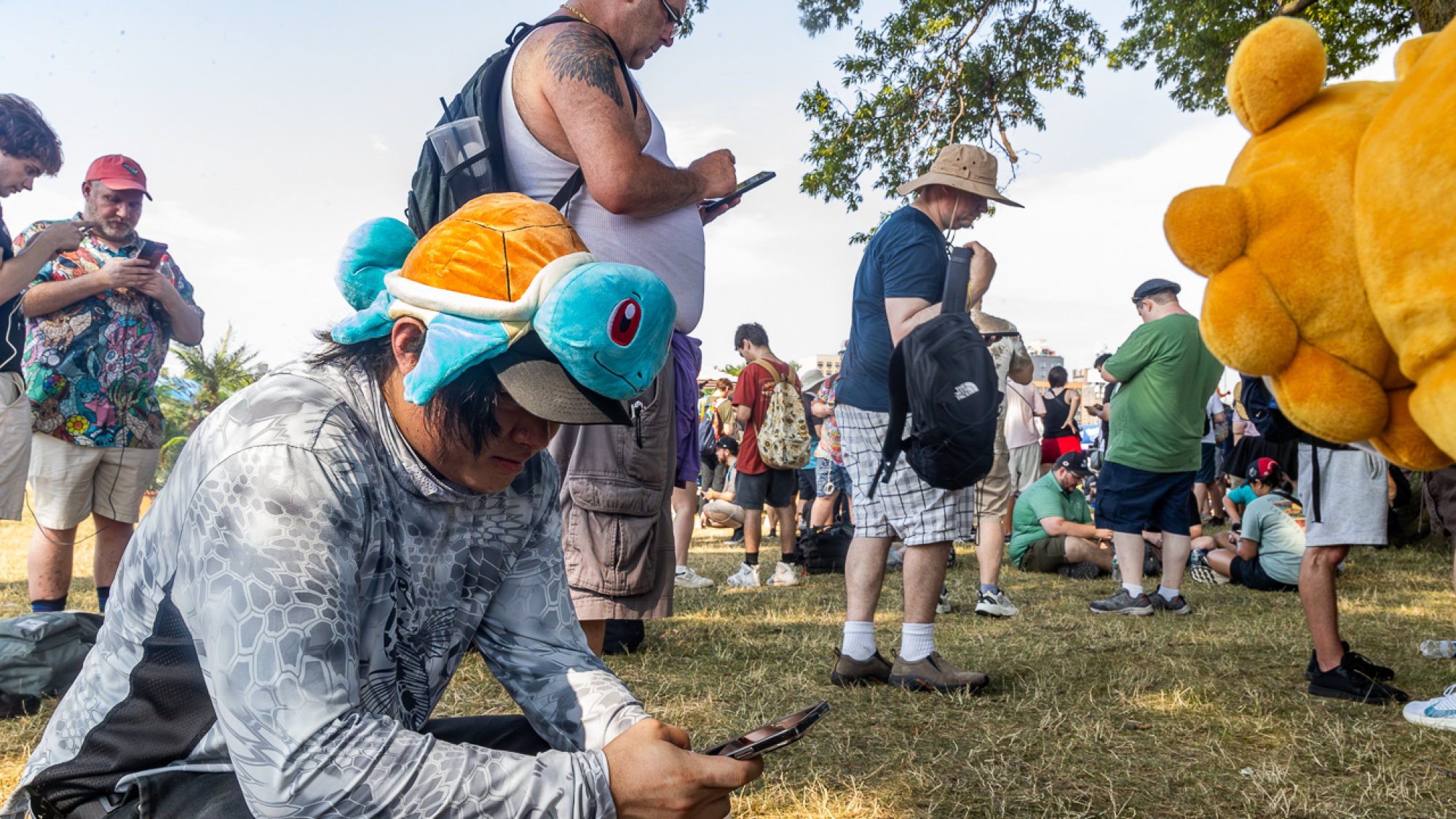 A man with a Squirtle hat squats on a lawn playing Pokémon GO.