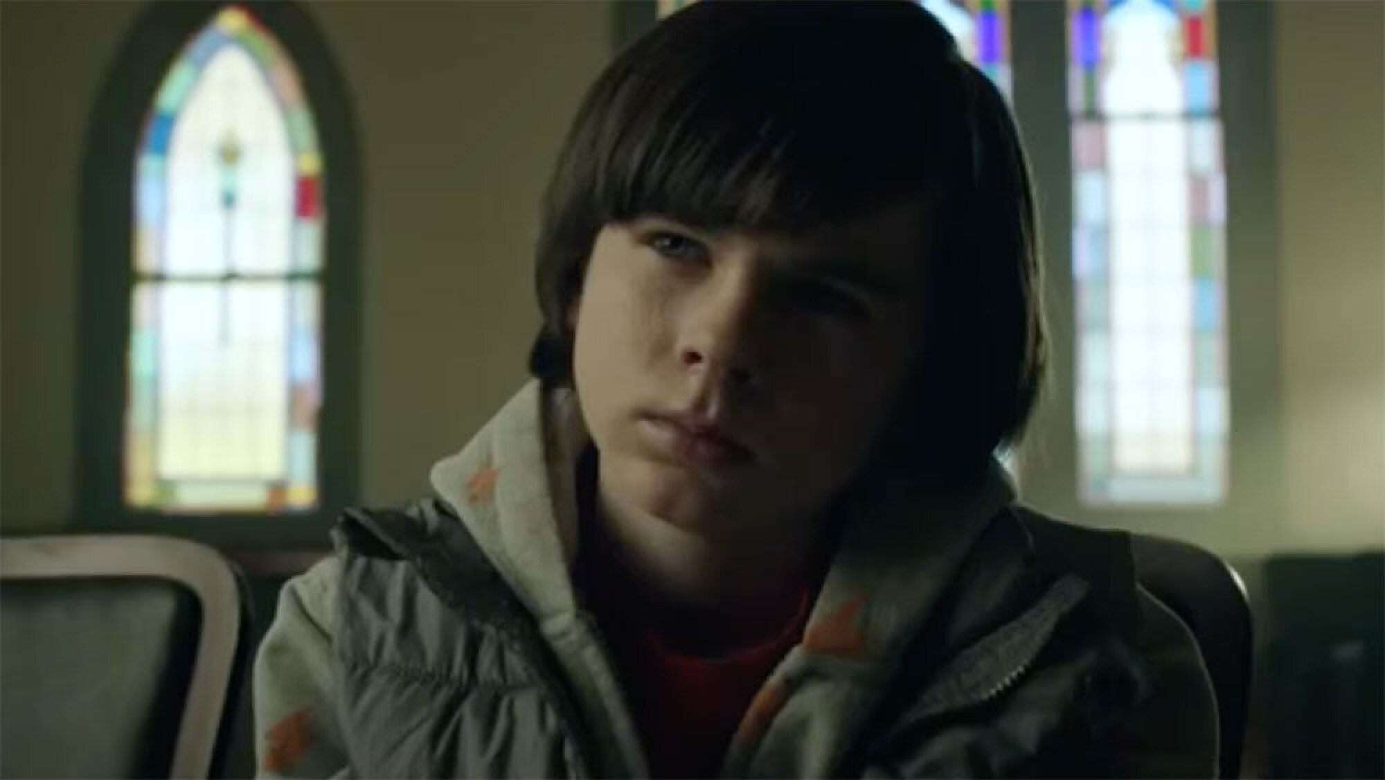 A boy sits in a church with stained glass windows behind him.