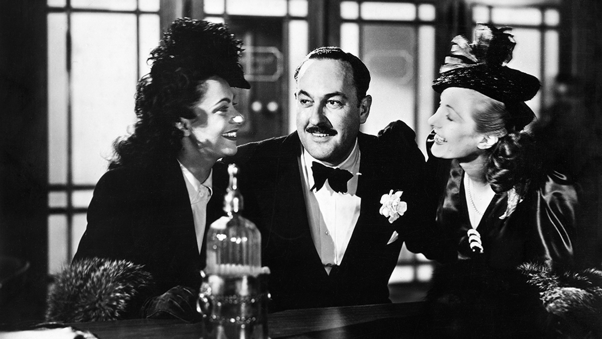 A grinning man sits in between two laughing women at a bar in a black-and-white image.