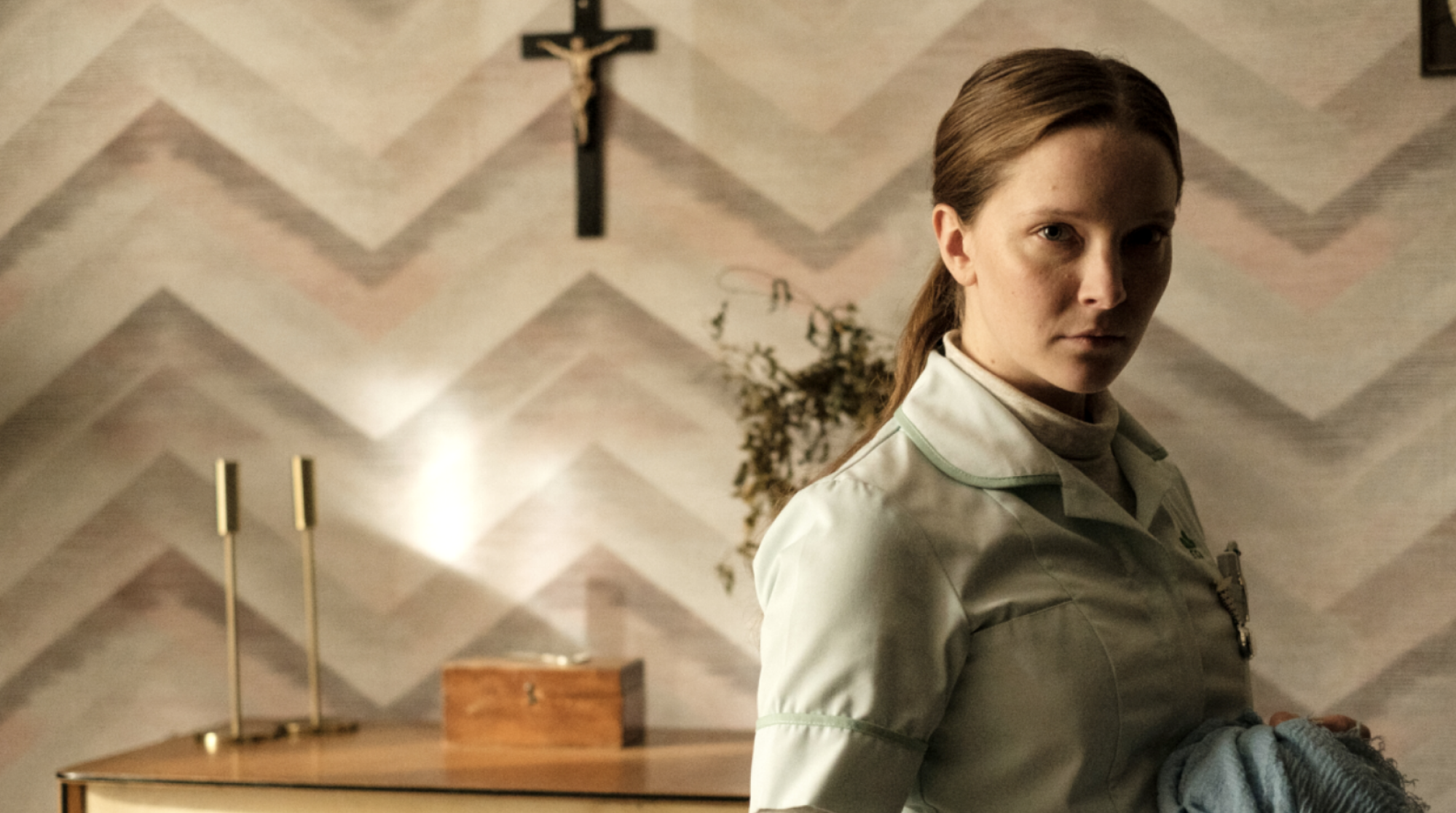 A woman stands in a room staring at the camera with a crucifix in the background.