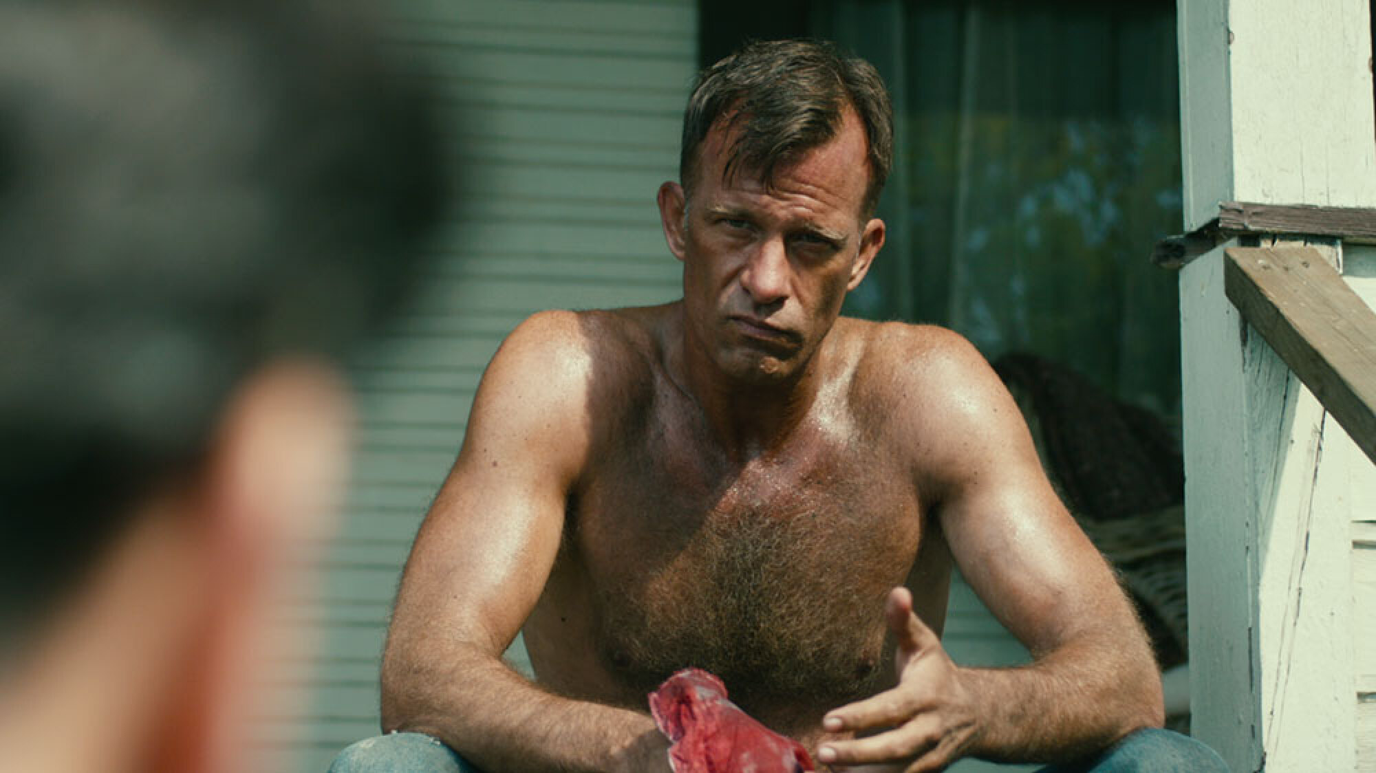 A shirtless man sitting on a porch looks mad.