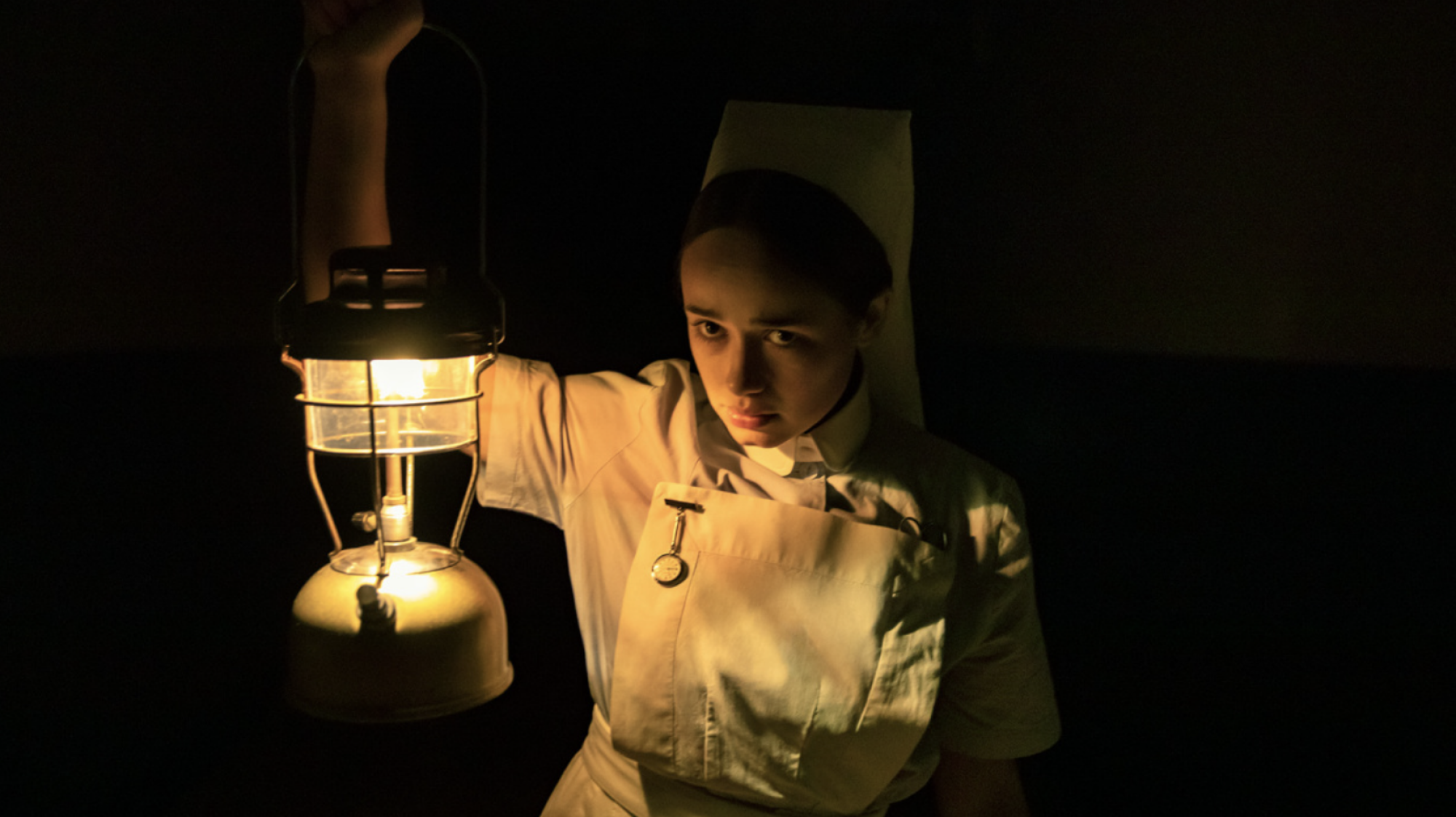 A woman in an old nurse's uniform stands in the dark holding a lantern.