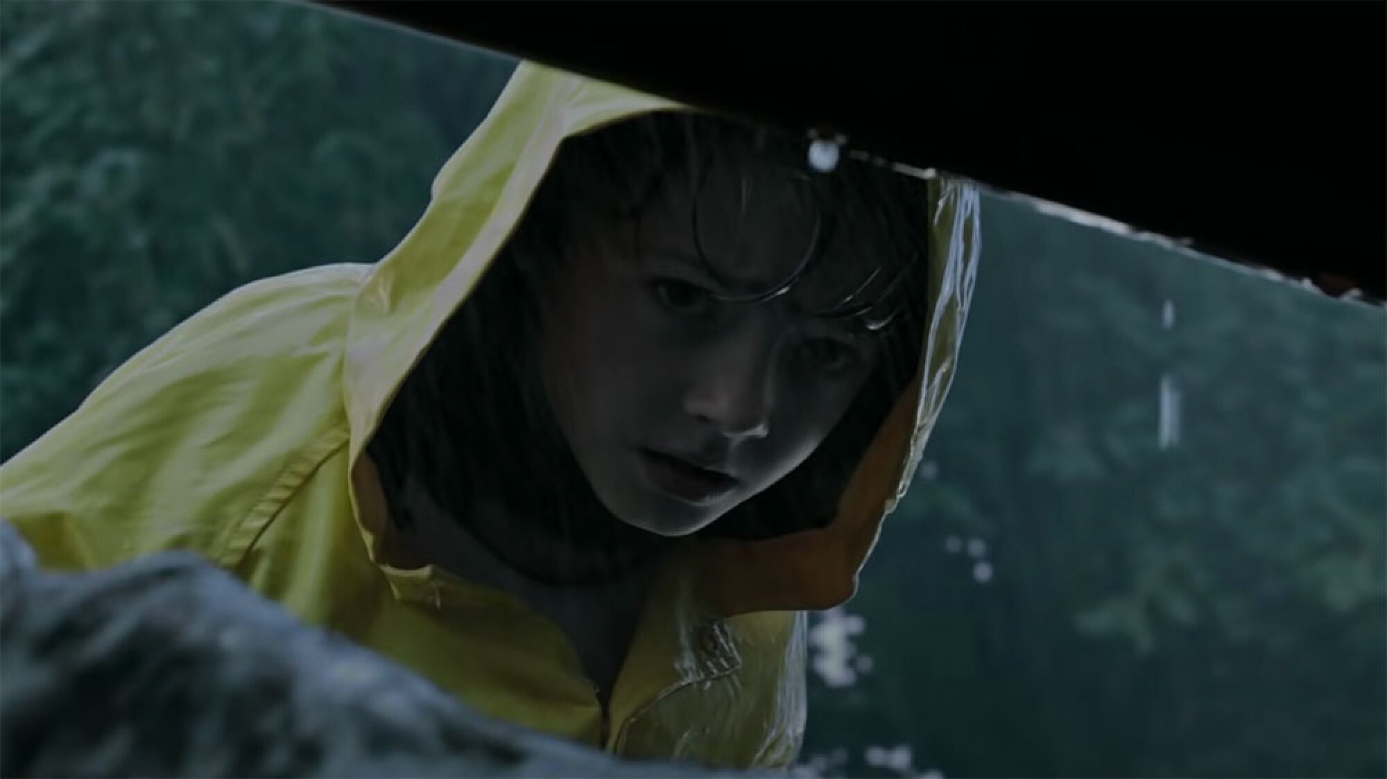 A boy in a yellow raincoat looks into a drain.